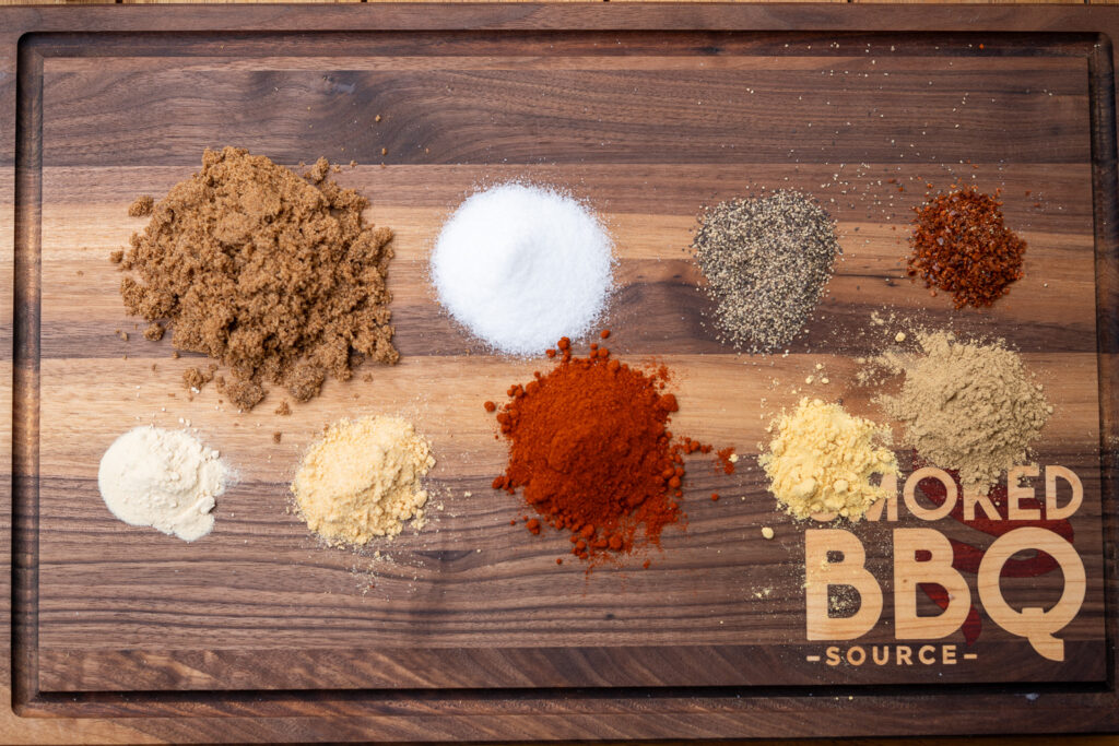 “Overhead view of pork rub ingredients arranged on a wooden cutting board, including brown sugar, salt, black pepper, paprika, garlic powder, onion powder, mustard powder, ground ginger, and chili flakes. Smoked BBQ Source logo engraved in the board.