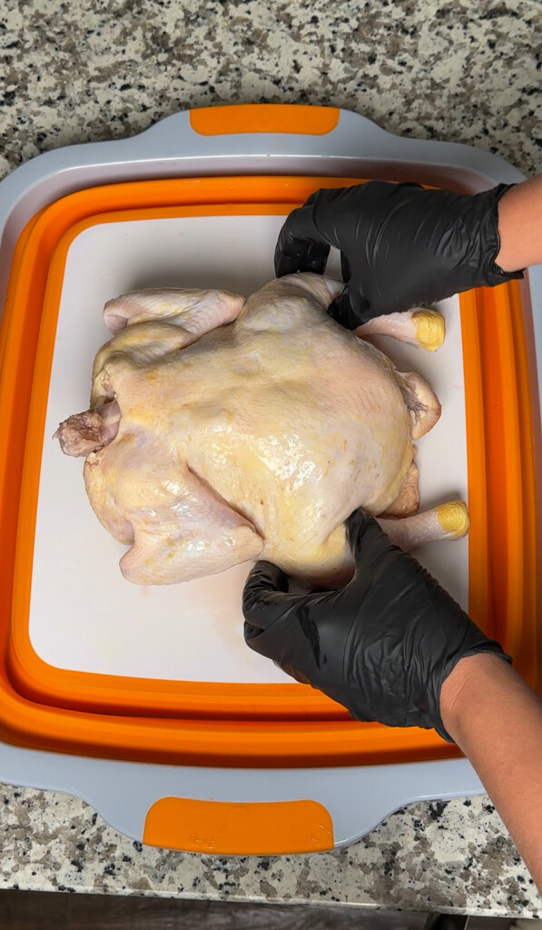 a raw chicken, backbone side up, on a white board, with black gloved hands either side