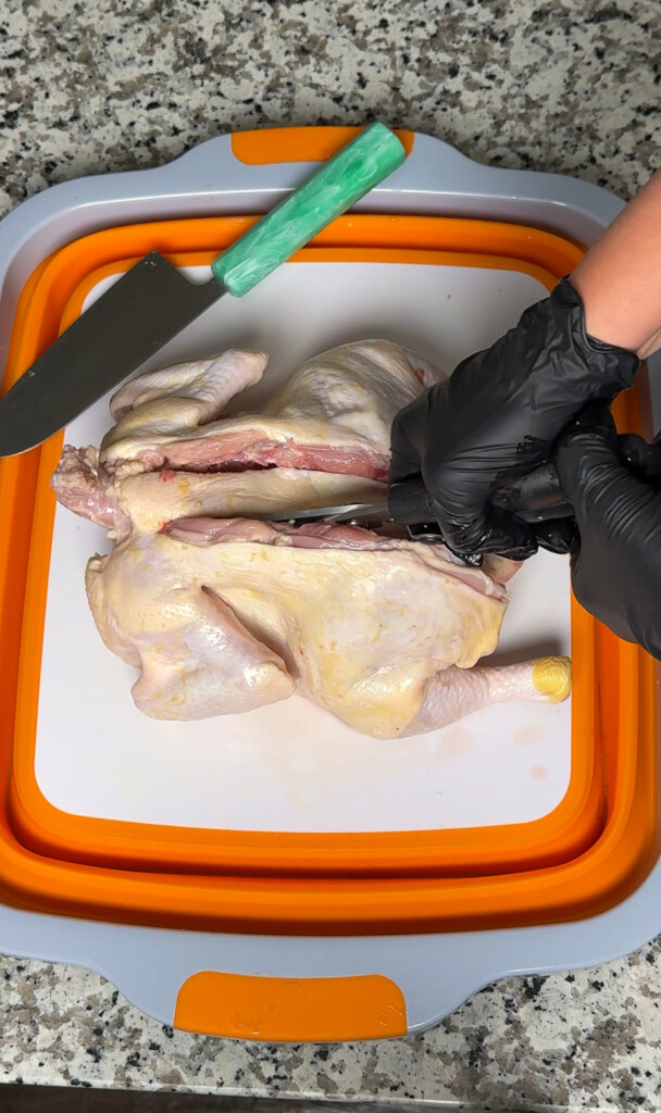 black gloved hands cutting the backbone out of a raw chicken using kitchen shears