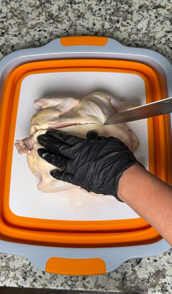 a knife slicing down the side of a raw chicken backbone, a black gloved hand holding the chicken