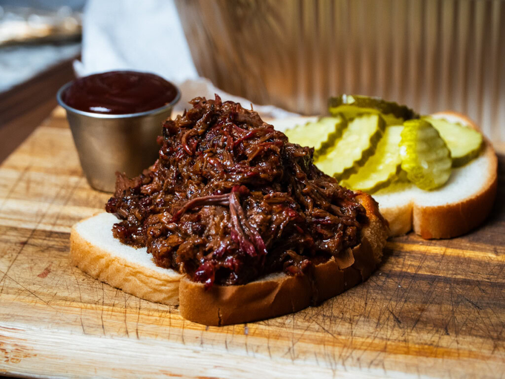 Pulled smoked chuck roast piled onto a slice of white bread, served open-faced with barbecue sauce and pickles on the side.