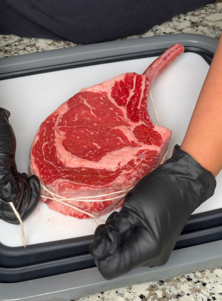 Close-up of a tomahawk steak being tied with butcher’s twine by gloved hands on a white cutting board, prepping it for even cooking.