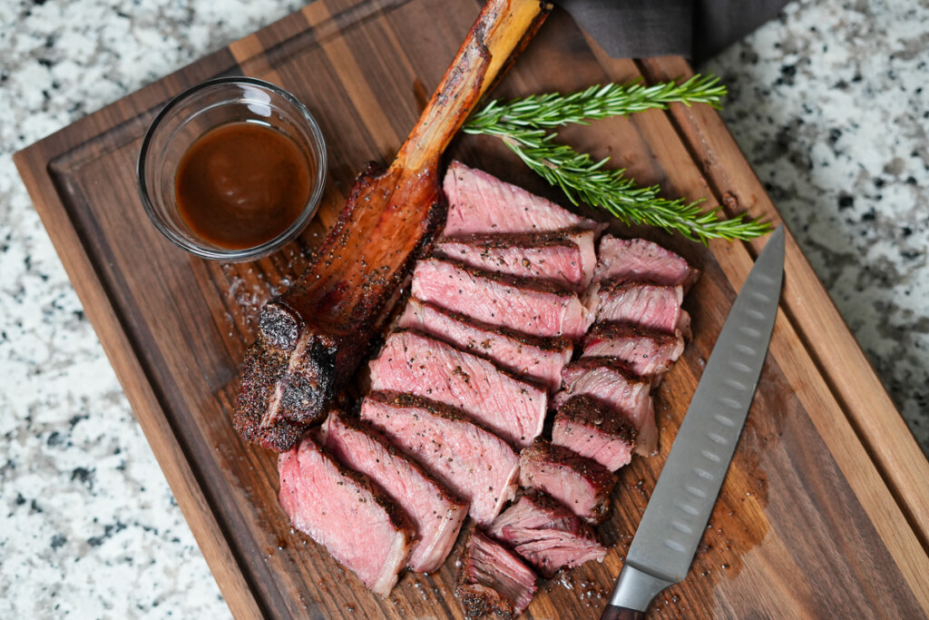 tomahawk steak sliced off the bone on a wooden board with a knife and some rosemary