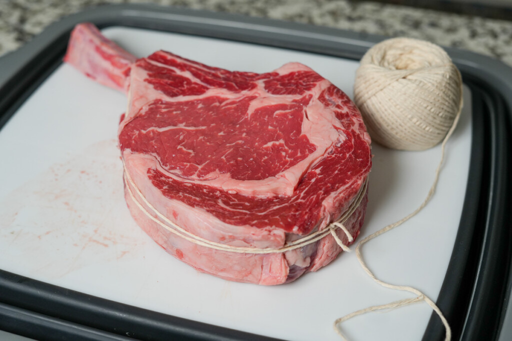 Raw tomahawk steak tied with butcher’s twine, resting on a cutting board with a spool of twine nearby, ready for seasoning and smoking.