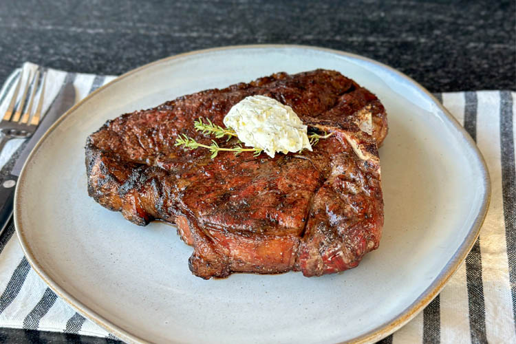 cooked porterhouse on a white plate with a garlic herb compound butter on it