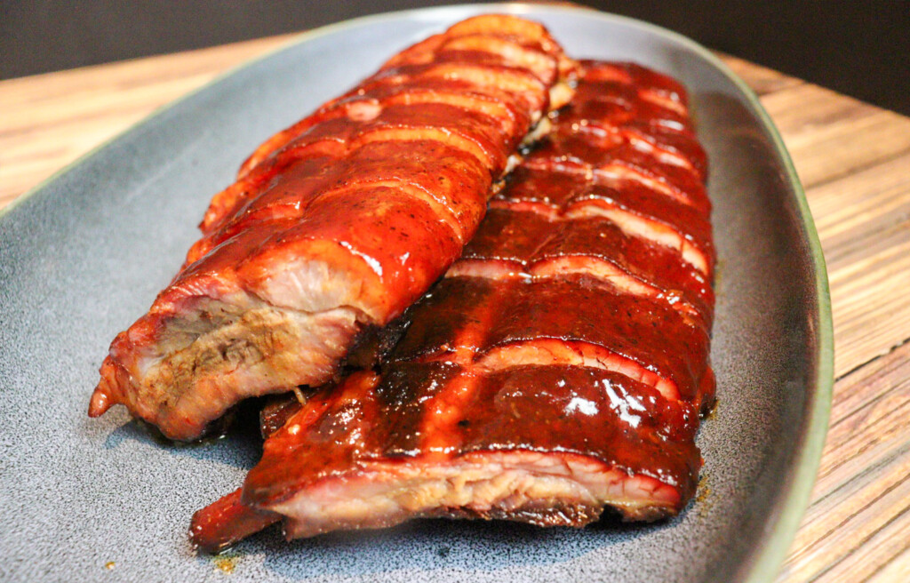 Hot and fast smoked baby back ribs glazed with BBQ sauce, sliced and served on a platter, showing a deep smoke ring, bark, and glossy finish.