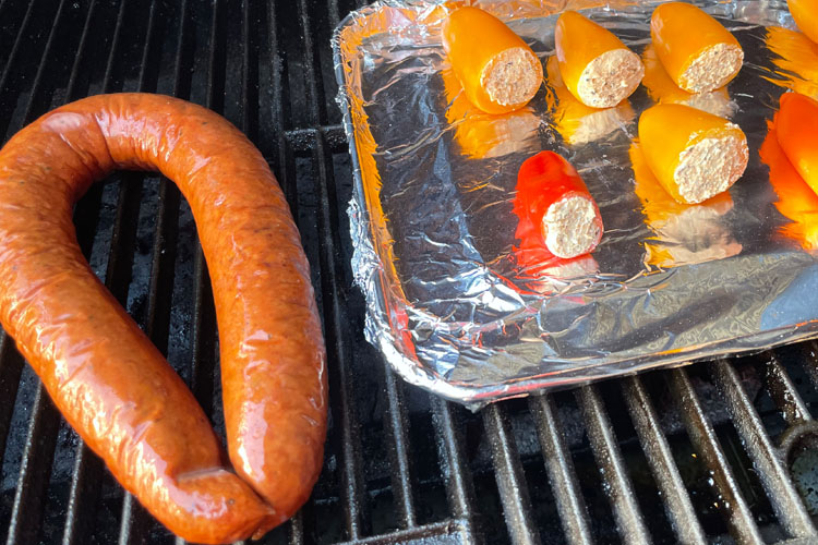 stuffed peppers on an aluminum foil covered tray, next to a sausage on the grill
