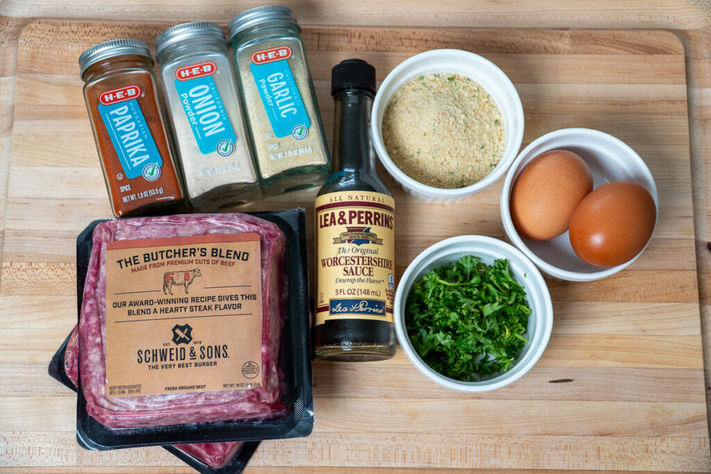 Ingredients for smoked barbecue meatballs, including ground beef, eggs, breadcrumbs, Worcestershire sauce, parsley, and seasonings like paprika, onion powder, and garlic powder, arranged on a wooden cutting board.