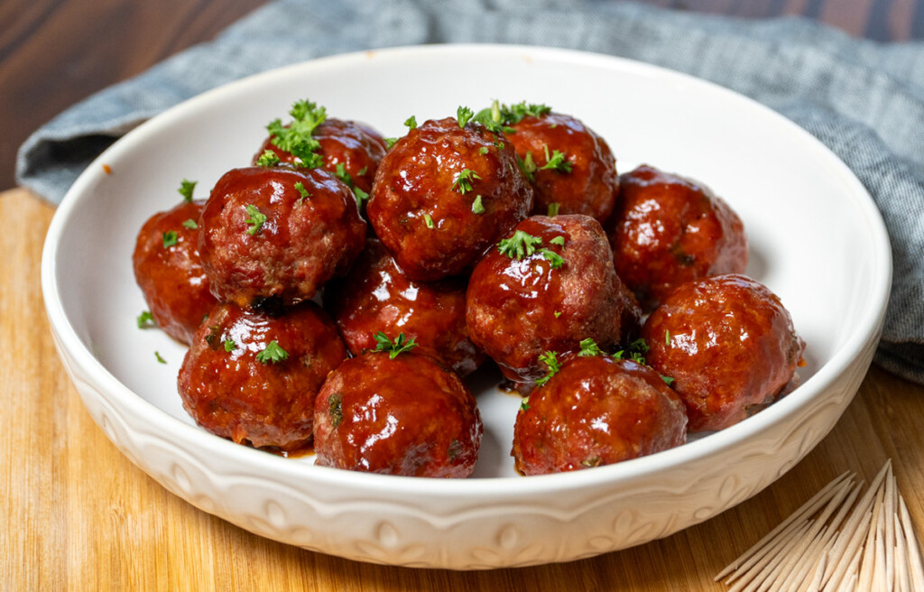 Smoked barbecue meatballs coated in a glossy, sweet, and smoky BBQ sauce, garnished with fresh parsley, served in a white bowl on a wooden board with toothpicks on the side.