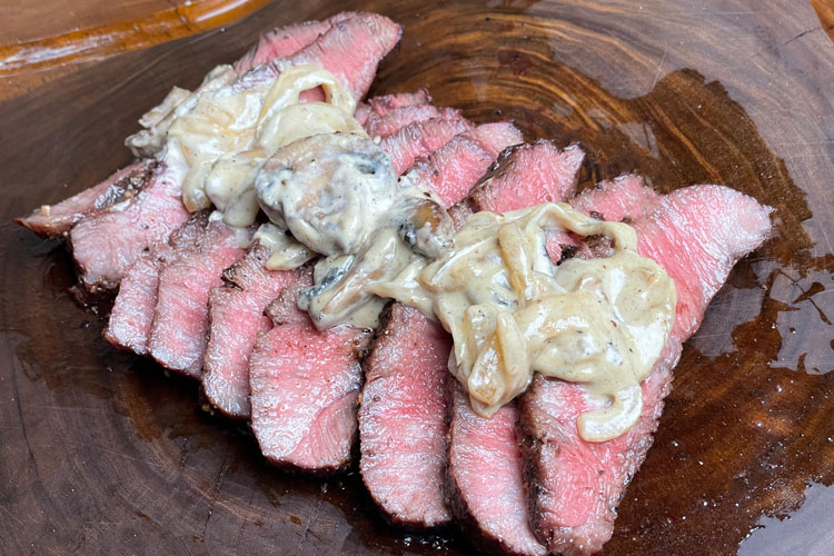 sliced flat iron steak with mushroom sauce on top on a wooden chopping board