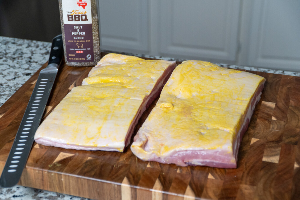 two pieces of raw pork belly with mustard smeared on them, sitting on a wooden board
