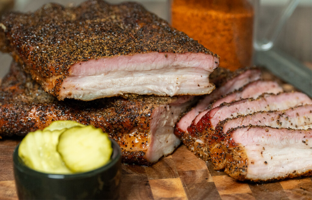 Overhead view of sliced smoked pork belly with dark bark and juicy pink interior on a wooden cutting board.