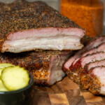 Overhead view of sliced smoked pork belly with dark bark and juicy pink interior on a wooden cutting board.