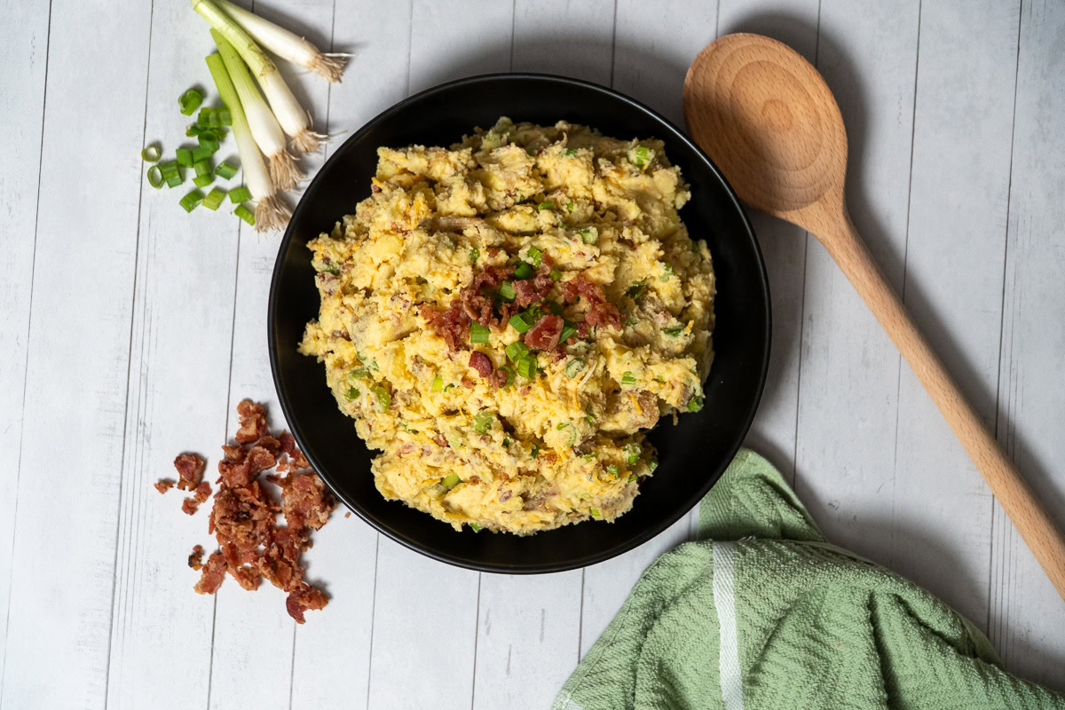 looking down on a bowl of loaded potato salad sarrounded by bacon, green onions, a wooden spoon and a green cloth
