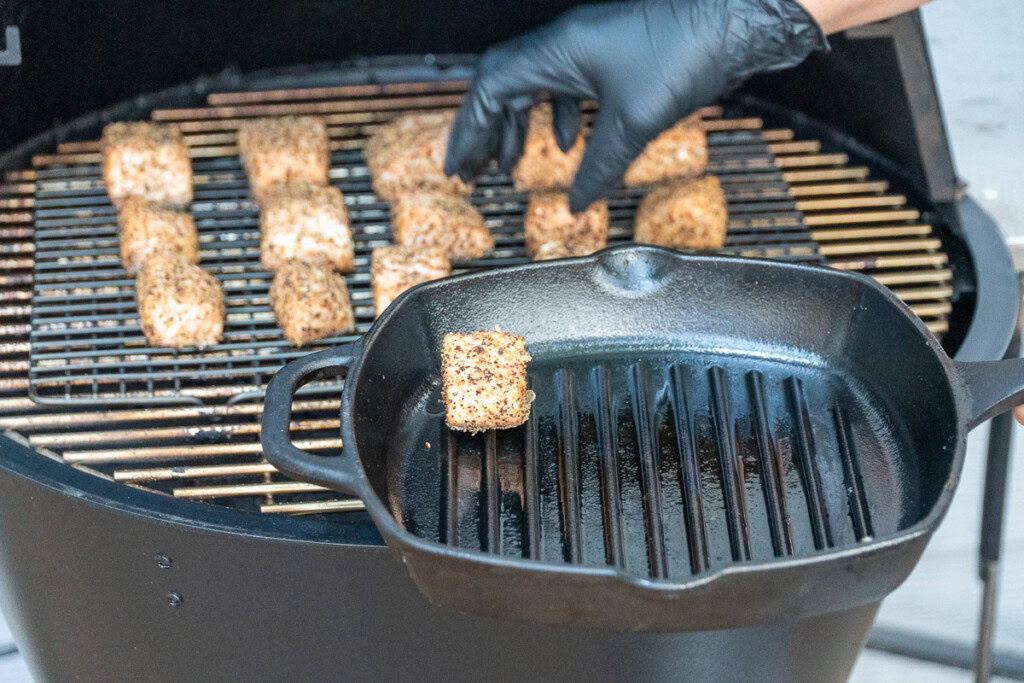 cubes of cooked salmon are being taken off the smoker and put in a cast iron pan