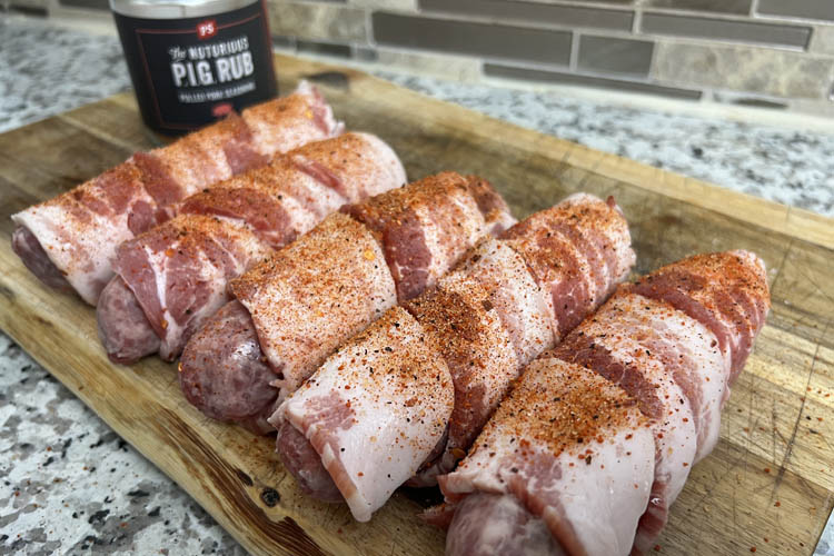 raw bacon wrapped brats, seasoned, on a wooden chopping board