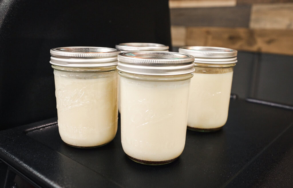Four glass mason jars filled with creamy white rendered beef tallow, arranged on a black grill shelf.