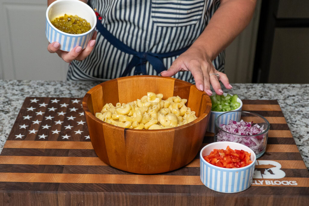cooked pasta in a wooden bowl with a person adding a bowl of pickle relish
