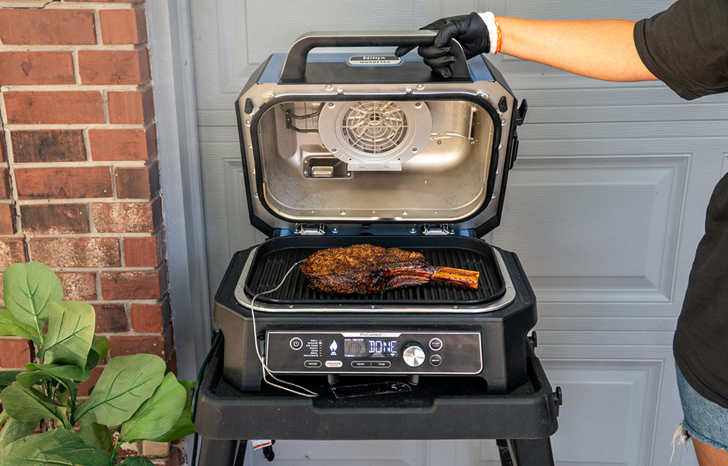 A cooked tomahawk steak rests on the grill grate of a Ninja Woodfire Pro XL, with the lid open to reveal the interior heating fan. A hand wearing a black glove holds the lid open, while the grill sits on a black cart in front of a garage door and red brick wall.