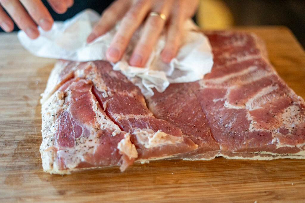 Hands patting cured pork belly dry with paper towels on a cutting board after rinsing.