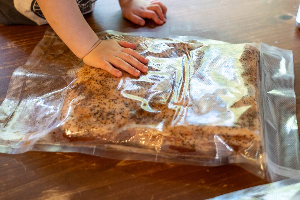 Child's hand pressing on vacuum-sealed slab of pork belly coated with pepper, part of the home bacon-curing process.