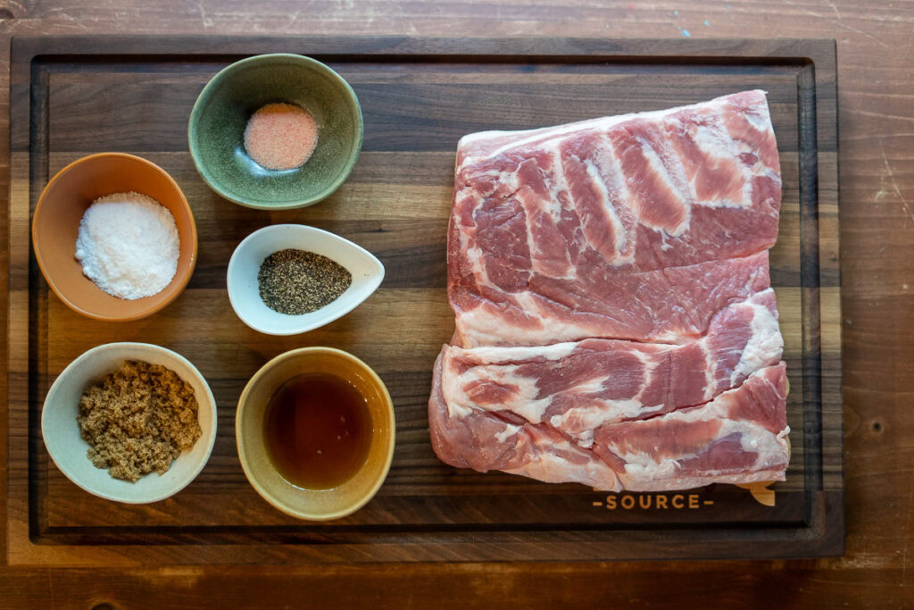 Uncured pork belly on a cutting board with bowls of curing salt, kosher salt, black pepper, brown sugar, and maple syrup arranged for homemade bacon preparation.