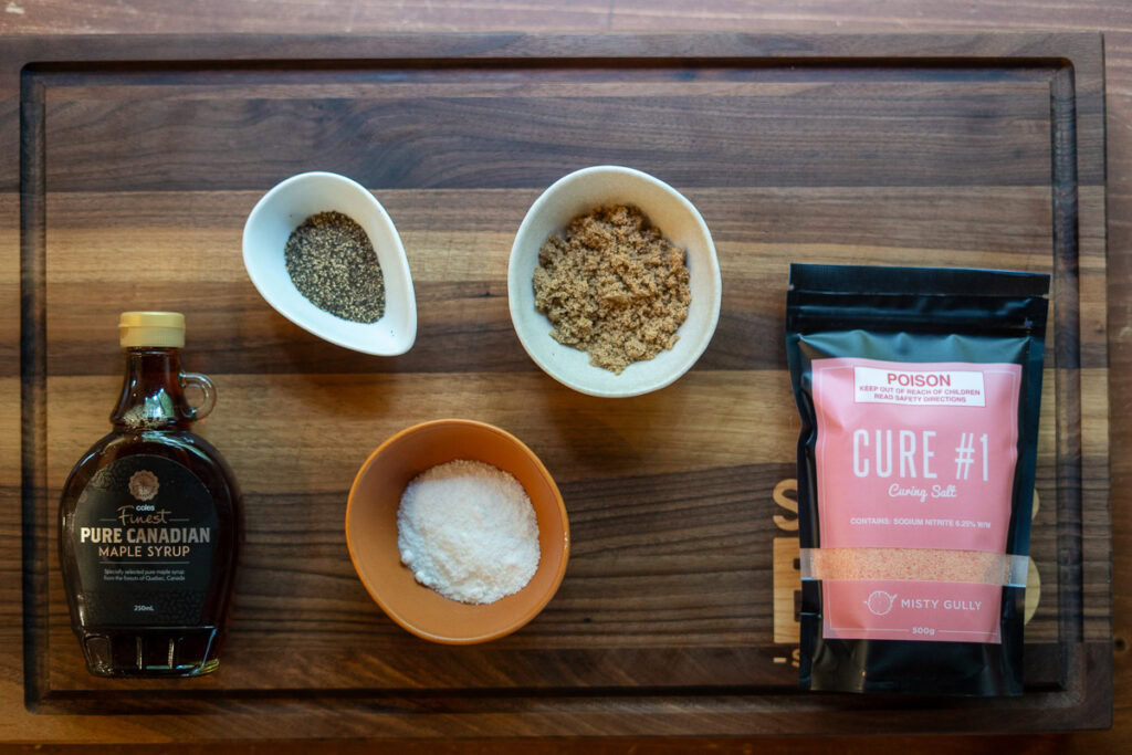 Ingredients for homemade bacon laid out on a cutting board, including maple syrup, kosher salt, brown sugar, black pepper, and a package of Cure #1 curing salt.