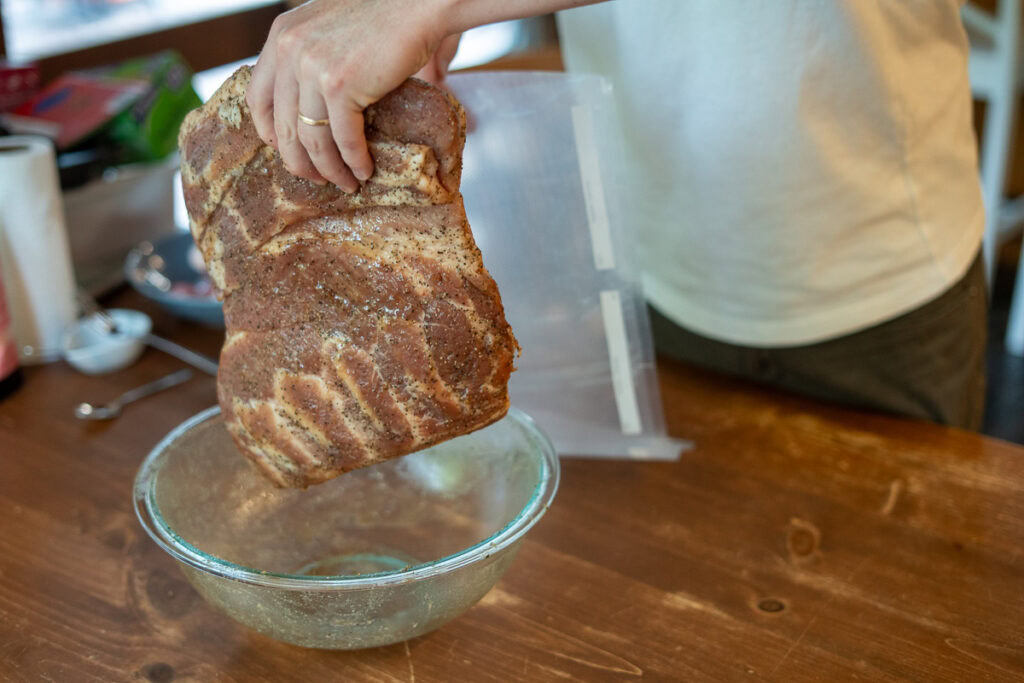 Hand placing a pork belly coated in dry cure into a vacuum seal bag after seasoning for homemade bacon preparation.