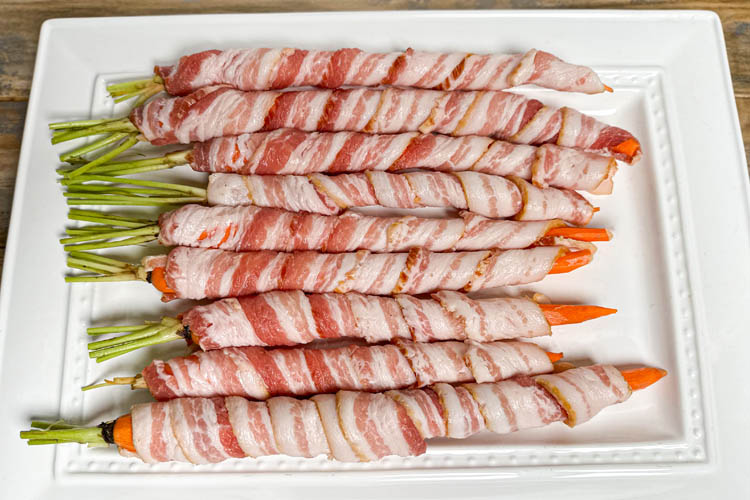 raw carrots wrapped in bacon on a white plate