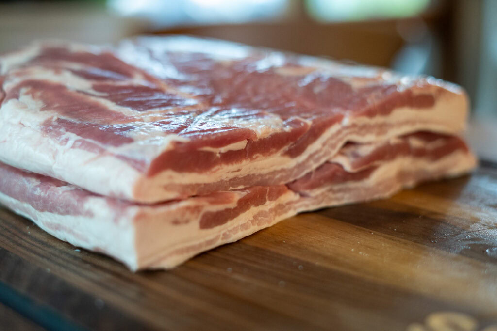 Close-up of raw pork belly slabs stacked on a wooden cutting board, showing layers of fat and meat before curing.