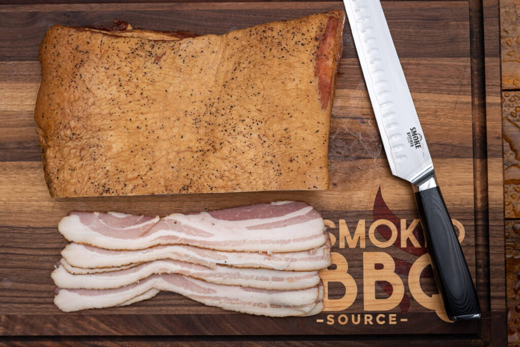 Slab of smoked homemade bacon with peppered crust shown alongside evenly sliced raw bacon strips on a wooden cutting board.