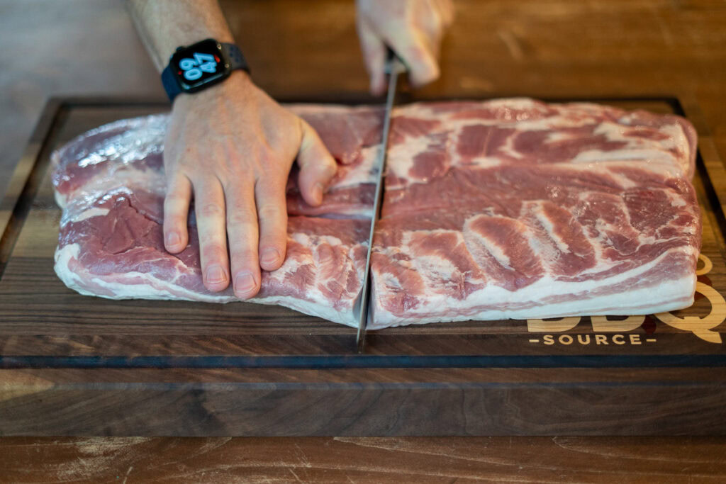 Person slicing a large slab of raw pork belly in half on a wooden cutting board, preparing it for curing.