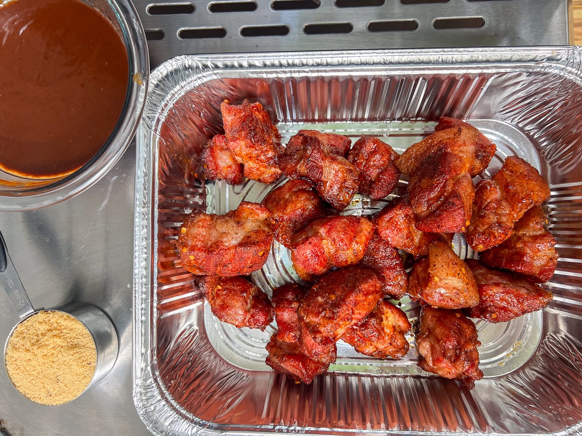 Pork burnt ends in an aluminum pan, coated in melted butter, brown sugar, and BBQ sauce.