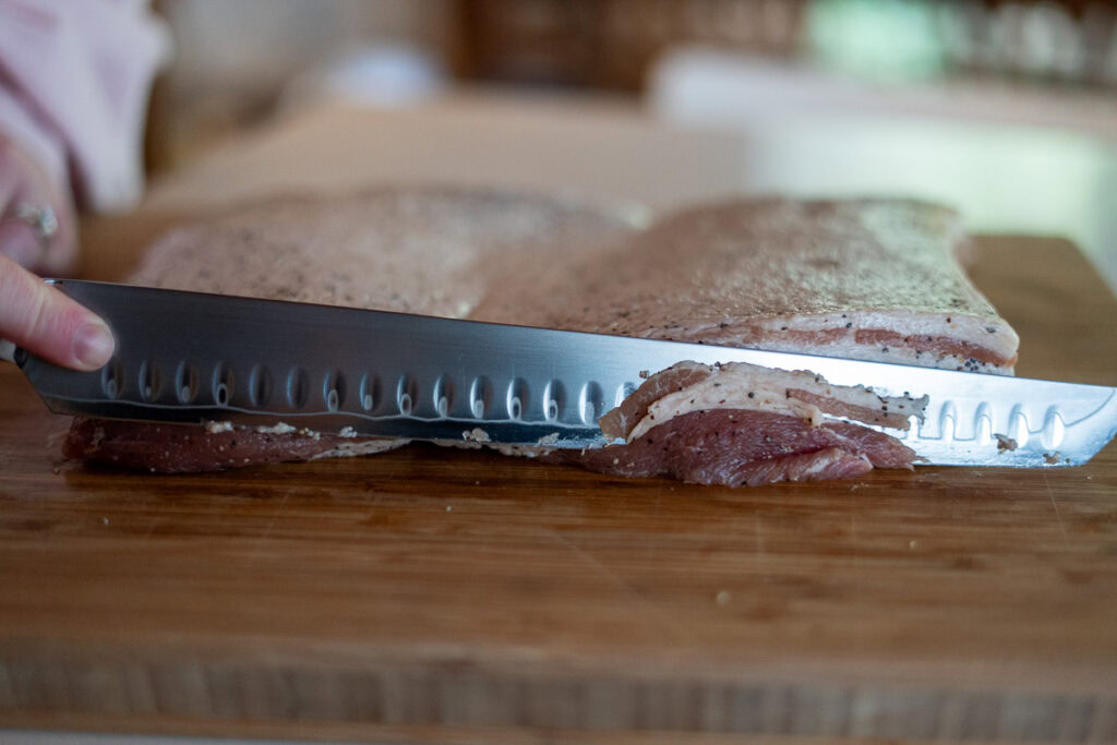 Knife slicing homemade cured bacon into thin strips on a wooden cutting board.