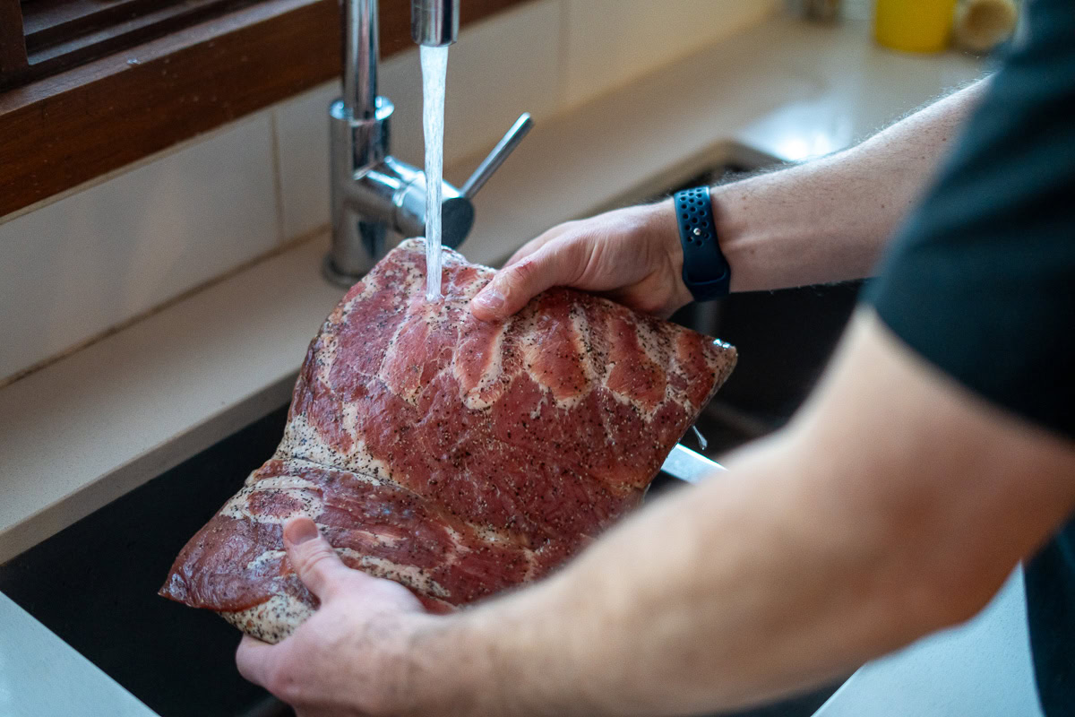 Person rinsing cured pork belly under cold running water in a kitchen sink to remove excess cure before smoking.