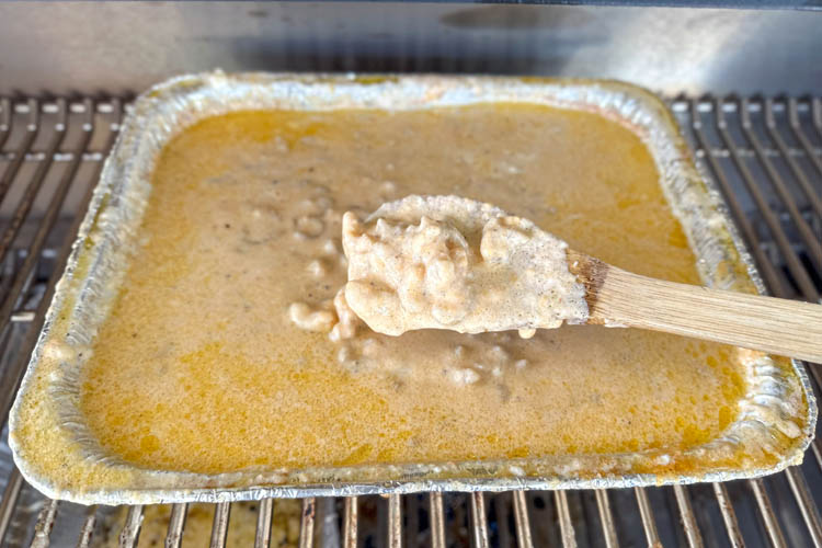 mac and cheese in tin tray on the grill being stirred with wooden spoon