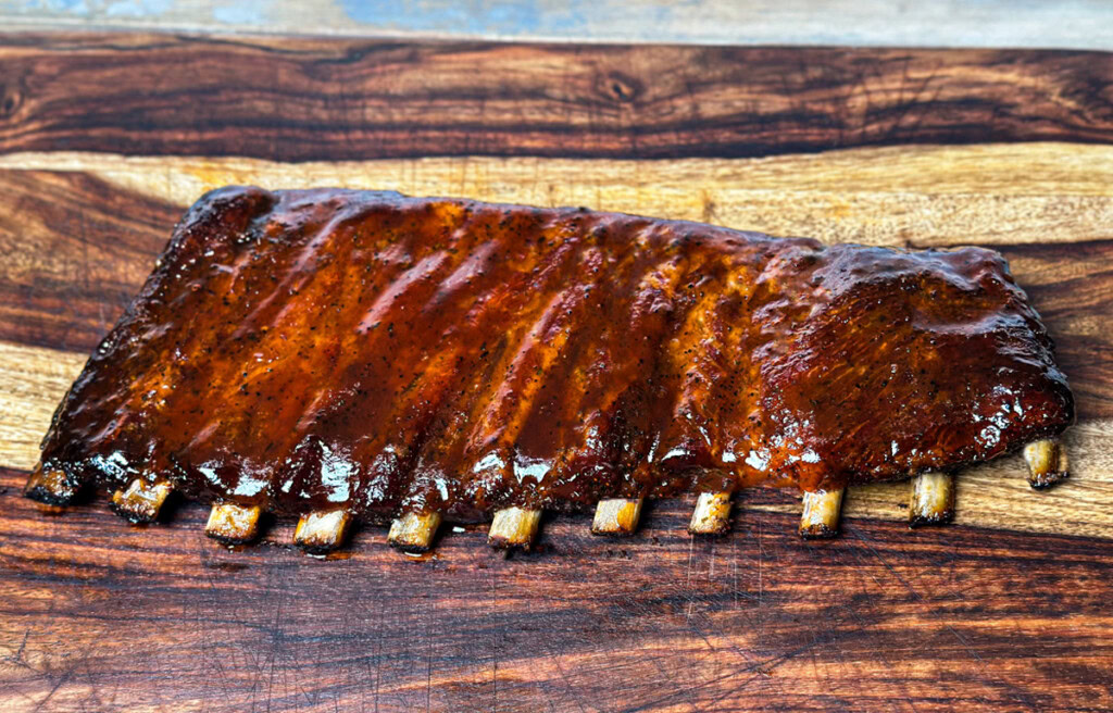 Glazed rack of St. Louis-style pork ribs on a wooden cutting board, cooked in an electric smoker with visible bone pullback and a caramelized BBQ sauce finish.
