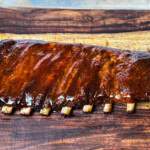 Glazed rack of St. Louis-style pork ribs on a wooden cutting board, cooked in an electric smoker with visible bone pullback and a caramelized BBQ sauce finish.