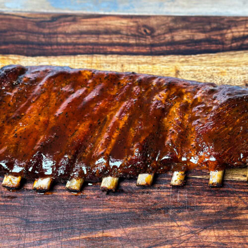 Glazed rack of St. Louis-style pork ribs on a wooden cutting board, cooked in an electric smoker with visible bone pullback and a caramelized BBQ sauce finish.