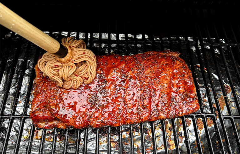 Close-up of ribs being basted with mop sauce on a smoker, using a traditional cotton mop brush. The meat is glistening with a rich, red glaze, surrounded by grill grates and foil-lined drip trays.