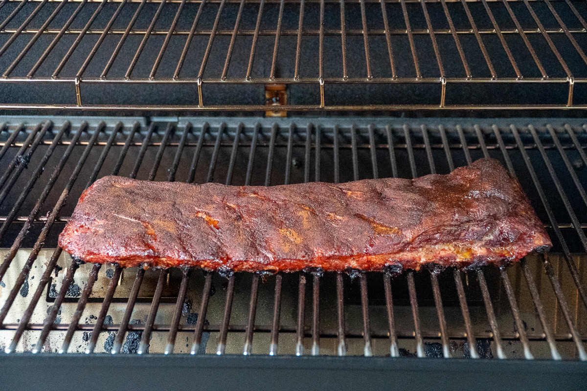 Rack of pork ribs on a smoker after three hours of smoking, showing darkened bark and surface caramelization.