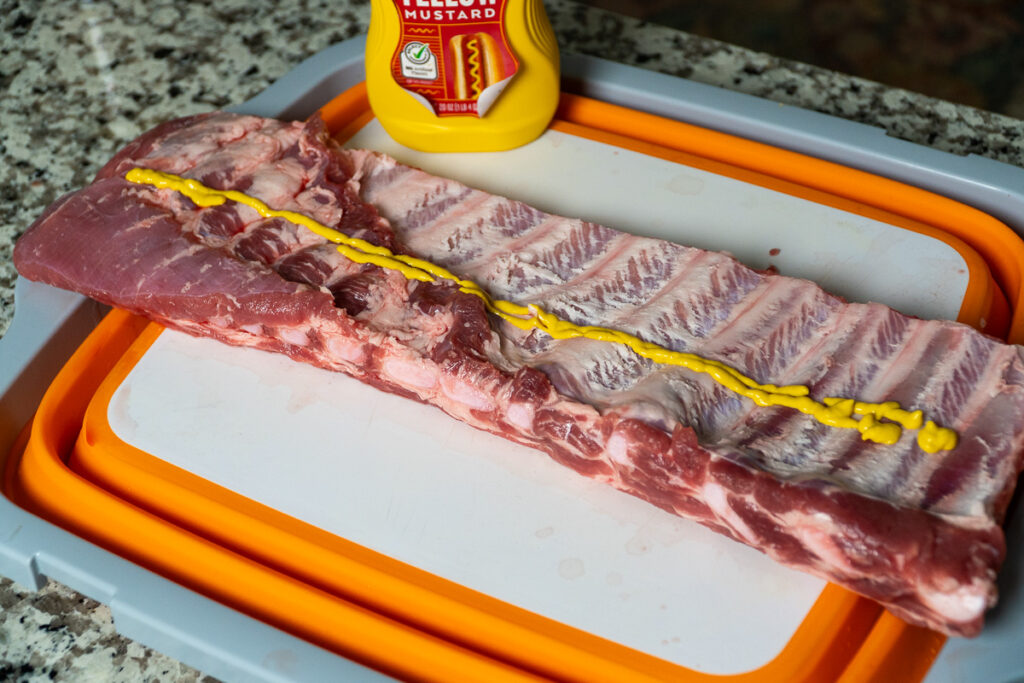 Raw rack of pork ribs on a cutting board with yellow mustard applied as a binder, ready for seasoning.