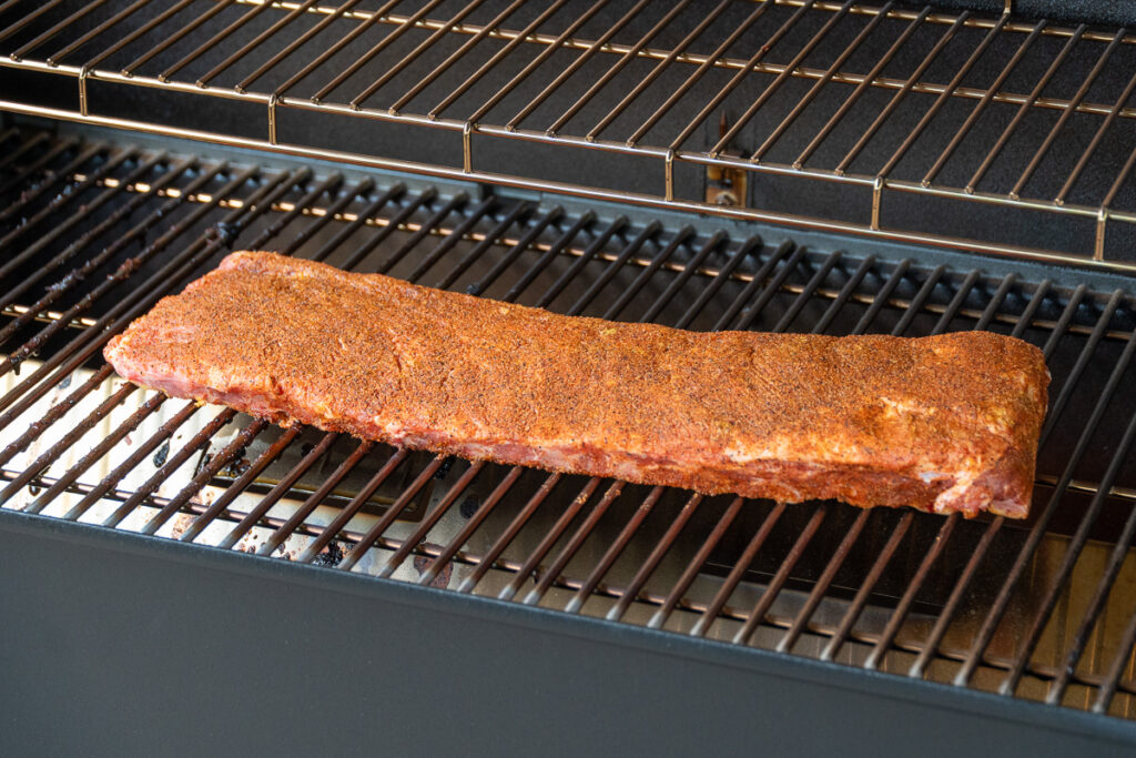 Rack of seasoned pork ribs placed meat-side up on a smoker grate during the first stage of the 3-2-1 method.
