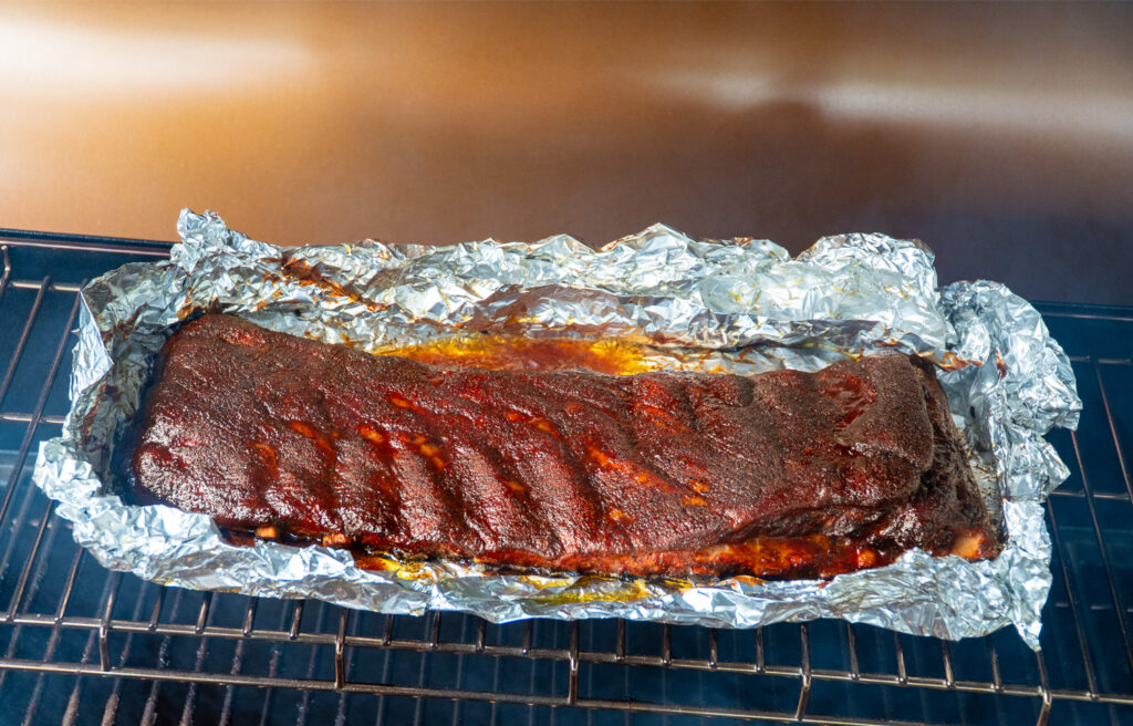 Glazed rack of smoked ribs resting in an open foil boat on the smoker, with sauce set and bark darkened during the final stage of the 3-2-1 method.