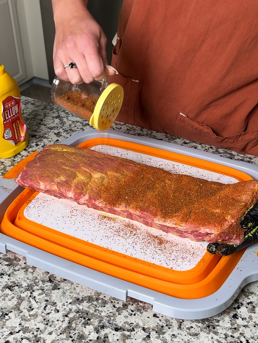 Person sprinkling dry rub over a rack of pork ribs coated with yellow mustard on a cutting board.