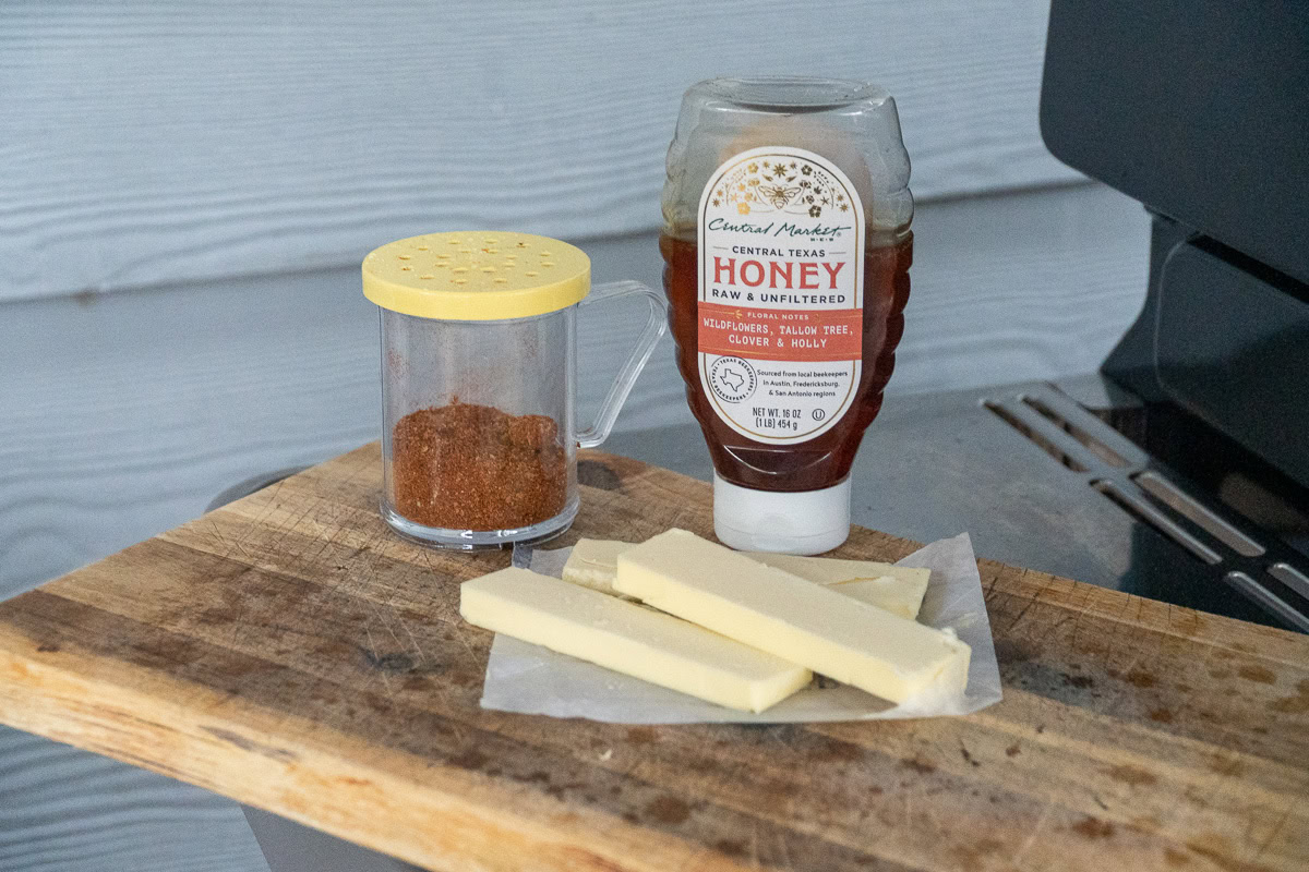 Butter, BBQ dry rub, and a bottle of Central Market honey on a wooden cutting board next to a grill — ingredients for wrapping smoked ribs.