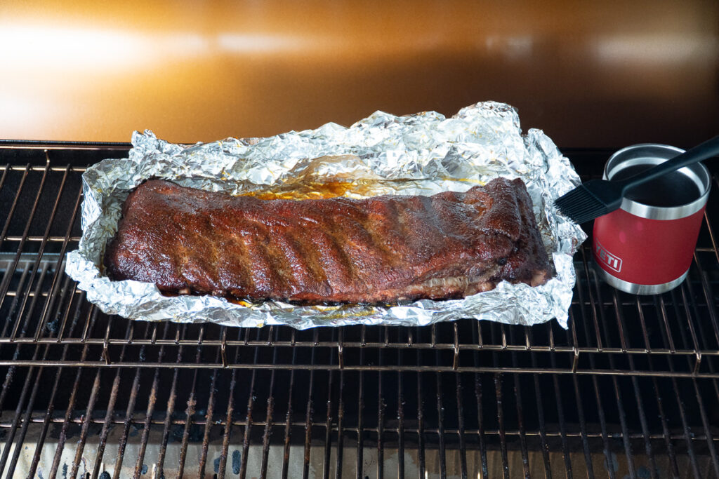 Unwrapped rack of ribs resting in foil on a smoker with a sauce pot and basting brush beside it, ready for the final glaze during the 3-2-1 method.