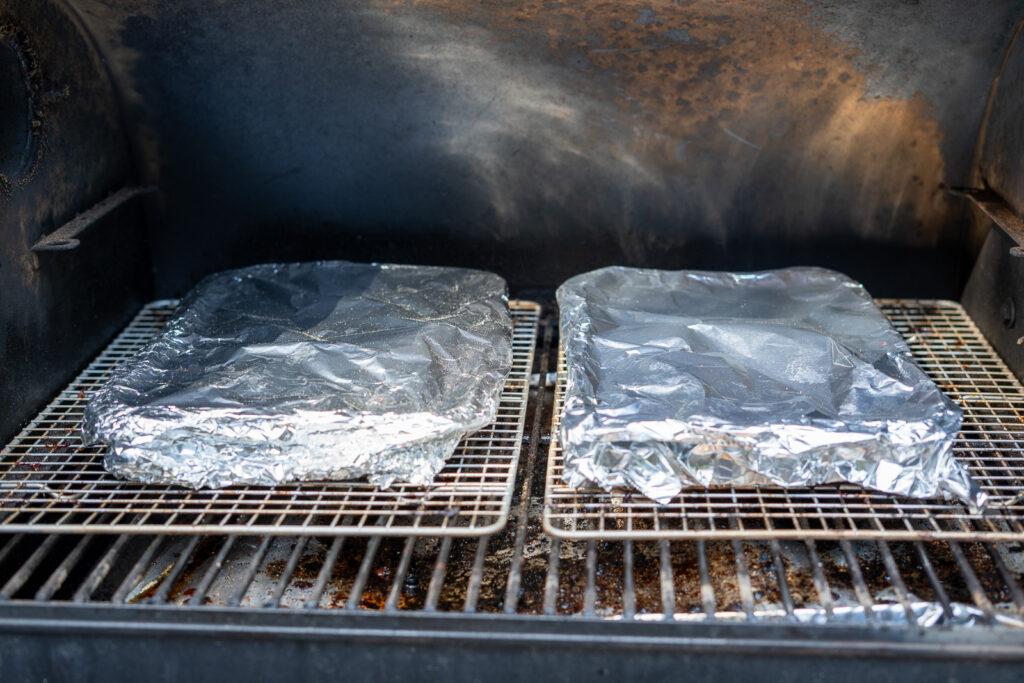 Two foil trays covered with aluminum foil inside a smoker, beginning the braising phase for pork belly burnt ends.