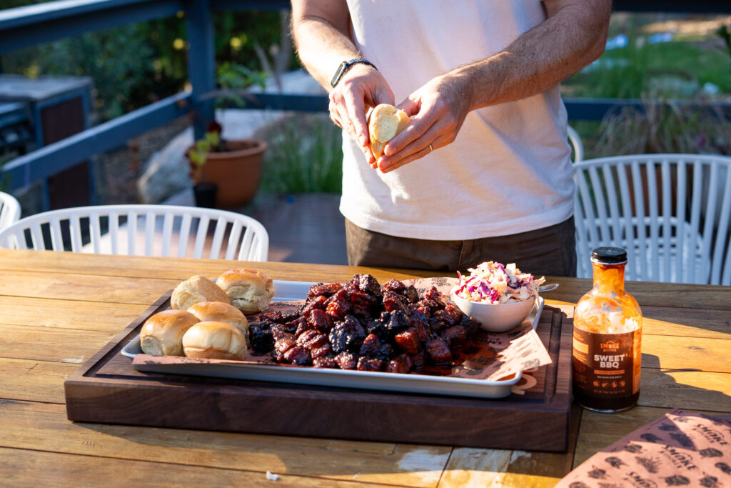 Person assembling pork belly burnt end sliders at an outdoor table, with a tray of glazed burnt ends, brioche rolls, coleslaw, and BBQ sauce in view.