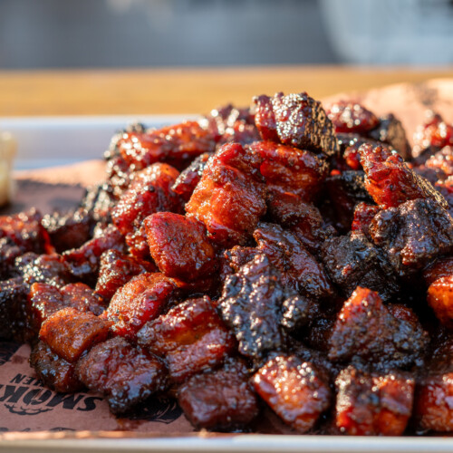 Close-up of glazed smoked pork belly burnt ends piled high on a tray with brioche buns and slaw, showcasing caramelized bark and sticky barbecue glaze.